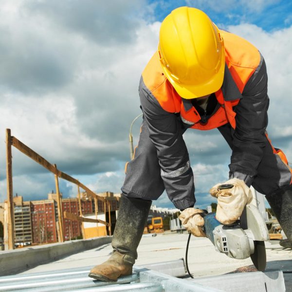 Construction worker using a power tool on a building site, wearing safety gear, with a cityscape in the background, related to RH Remodeling's expertise in public building renovations.