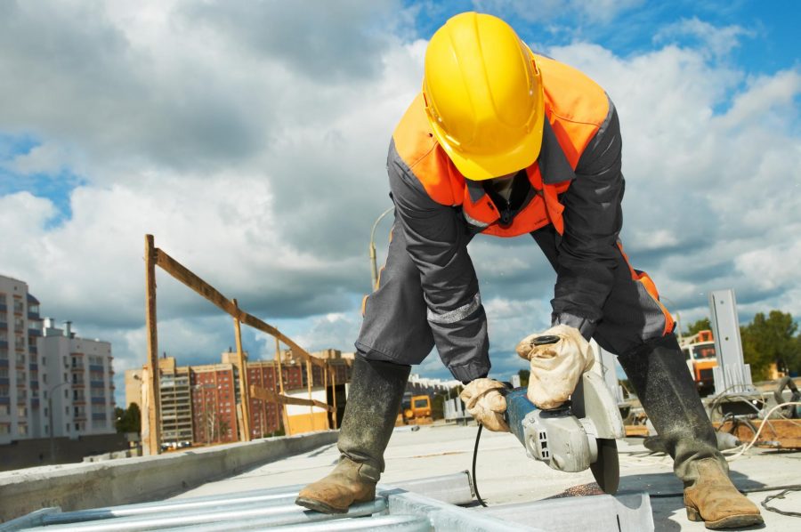 Construction worker using a circular saw on a building renovation site, wearing a hard hat and safety gear, with urban backdrop and cloudy sky.